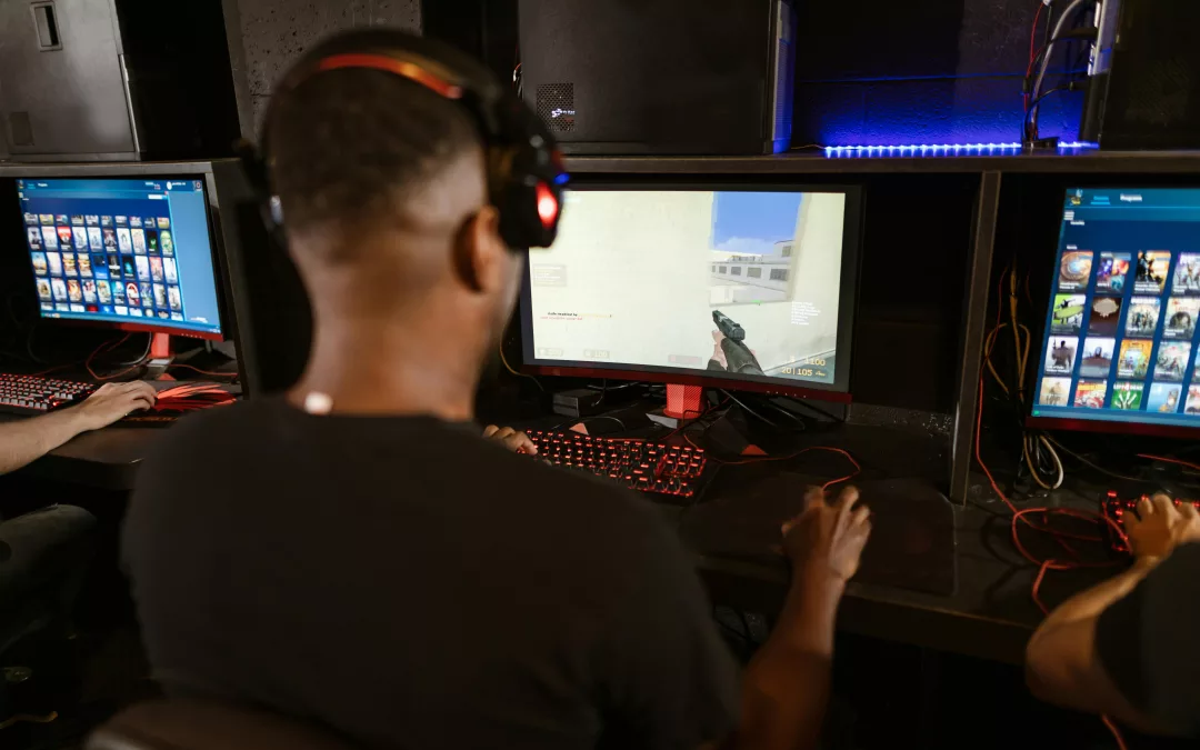 Un homme vu de dos, portant un casque audio, joue à un jeu de tir à la première personne sur un ordinateur dans une salle d’e-sport équipée de plusieurs stations de jeu.