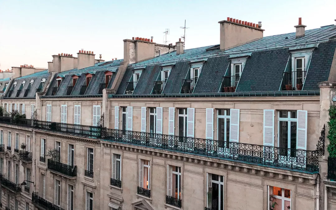 Vue oblique sur une rangée d’immeubles haussmanniens typiques : façades en pierre claire, balcons en fer forgé, toits mansardés en ardoise ponctués de cheminées alignées, sous un ciel bleu limpide en fin d’après-midi, avec une rue étroite en contrebas.