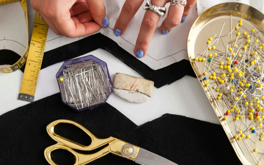 Atelier de couture avec tissu noir, ciseaux dorés, épingles et mains créant un vêtement artisanal durable.