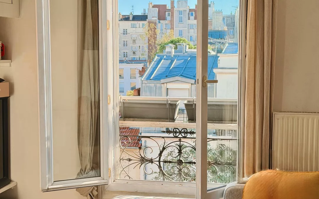 Vue d’un salon parisien baigné de lumière, avec une fenêtre ouverte donnant sur les toits et un canapé clair